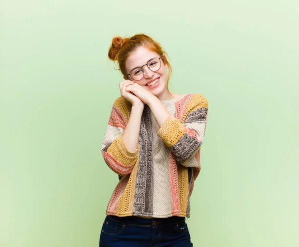 young pretty red head woman feeling in love and looking cute, adorable and happy, smiling romantically with hands next to face against green wall