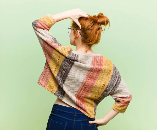 young pretty red head woman feeling clueless and confused, thinking a solution, with hand on hip and other on head, rear view against green wall