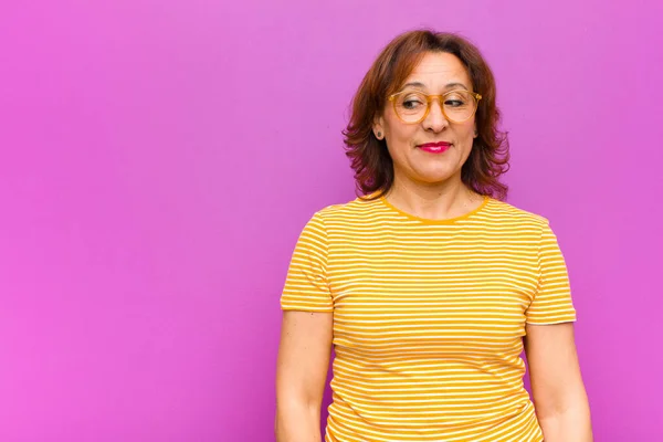 middle age woman wondering, thinking happy thoughts and ideas, daydreaming, looking to copy space on side against purple wall