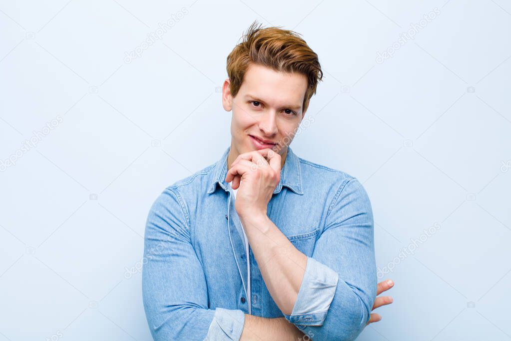 young red head man looking happy and smiling with hand on chin, wondering or asking a question, comparing options against blue wall