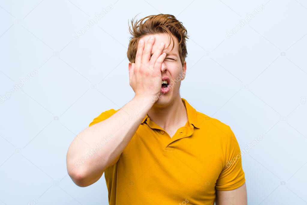 young red head man looking sleepy, bored and yawning, with a headache and one hand covering half the face against blue wall