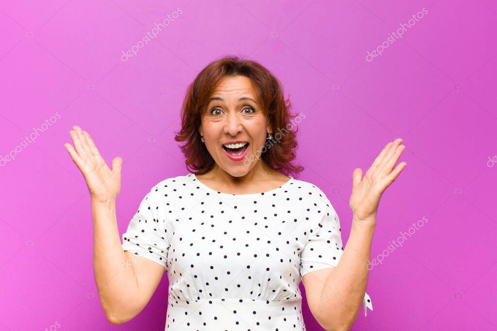 middle age woman feeling happy, excited, surprised or shocked, smiling and astonished at something unbelievable against purple wall