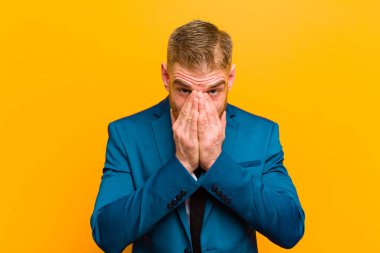 Genç kızıl saçlı işadamı mutlu, neşeli, şanslı ve şaşkın görünüyor. İki eli turuncu arka planda olan ağzını kapatıyor.