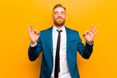 Genç kızıl saçlı işadamı konsantre ve meditasyon yapıyor gibi görünüyor, tatmin olmuş ve rahatlamış hissediyor, düşünüyor ya da turuncu arka plana karşı bir seçim yapıyor.