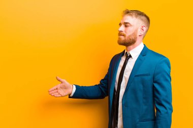 Genç kızıl saçlı işadamı gülümsüyor, sizi selamlıyor ve başarılı bir anlaşma için el sıkışma teklif ediyor, turuncu arka plana karşı işbirliği kavramı