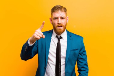 Genç kızıl saçlı işadamı kızgın agresif bir ifadeyle kameraya bakıyor turuncu arka planda kızgın, çılgın bir patron gibi görünüyor.