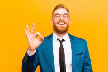 Genç kızıl saçlı işadamı kendini başarılı ve tatmin olmuş hissediyor. Ağzı açık gülümsüyor. Elleri turuncu arka planda.