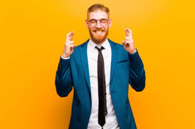 Genç kızıl saçlı işadamı gülümsüyor ve endişeyle iki parmağını çapraz yapıyor. Endişeli hissediyor ve turuncu arka planda iyi şanslar diliyor veya umut ediyor.