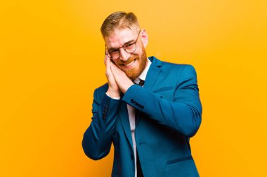 Genç kızıl saçlı iş adamı aşık ve sevimli görünüyor, sevimli ve mutlu, elleri turuncu arka planda, romantik bir şekilde gülümsüyor.