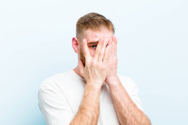 Genç kızıl saçlı adam elleriyle yüzünü kapatıyor, parmaklarının arasından şaşkın bir ifadeyle bakıyor ve yumuşak mavi arka plana bakıyor.
