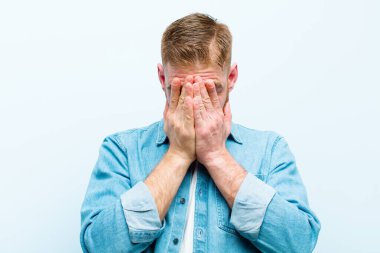 Genç kızıl saçlı adam üzgün, hayal kırıklığına uğramış, gergin ve depresif, iki eliyle yüzünü kaplayan, yumuşak mavi arka planda ağlayan
