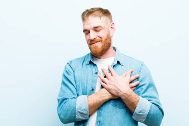 Genç kızıl saçlı adam üzgün, yaralı ve kalbi kırık görünüyor, iki elini de kalbe yakın tutuyor, ağlıyor ve mavi arka planda depresif hissediyor.