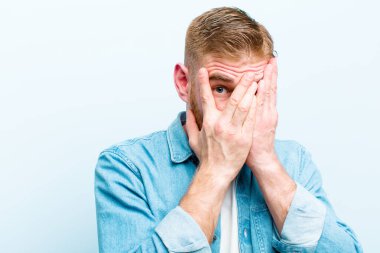 Genç kızıl saçlı adam korkmuş ya da utanmış hissediyor. Gözleri yarı kapalı, mavi arka planda dikizliyor ya da dikizliyor.