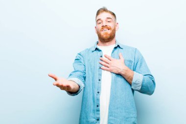 Genç kızıl saçlı adam mutlu ve aşık, bir eli kalbinin yanında gülümsüyor ve diğeri de yumuşak mavi arka plana doğru uzanıyor.