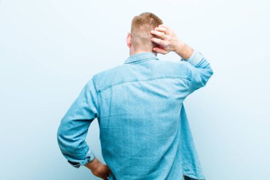 Genç kızıl saçlı adam bilgisiz ve kafası karışık hissediyor, bir çözüm düşünüyor, eli kalçasında, diğeri kafasında, arka planı yumuşak mavi.