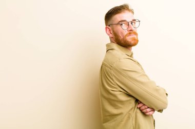 Genç kızıl saçlı adam neşeyle gülümsüyor, mutlu, tatmin olmuş ve rahatlamış hissediyor, kollarını kavuşturup bej arka plana bakıyor.