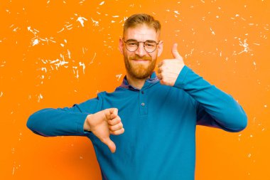 Genç kızıl saçlı adam kafası karışık, bilgisiz ve kararsız hissediyor, iyi ve kötü seçenekleri tartıyor veya grunge turuncu duvarına karşı seçenekleri değerlendiriyor.