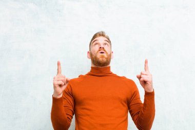 Kaplumbağa boynu takan genç kızıl saçlı adam şok olmuş, şaşkın ve ağzı açık, iki eliyle beton duvara uzayı işaret ediyor.