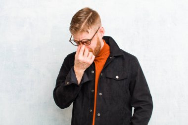 Genç kızıl saçlı adam stresli, mutsuz ve hayal kırıklığına uğramış, alnına dokunuyor ve grunge duvarına karşı şiddetli baş ağrısı çekiyor.