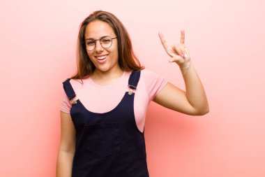 Genç kadın kendini mutlu, eğlenceli, kendinden emin, pozitif ve asi hissediyor, pembe arka planda kaya ya da heavy metal işareti yapıyor.