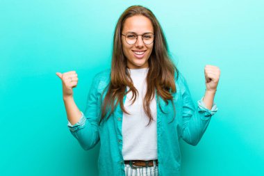 Neşeli gülümseyen, mutlu görünen, kaygısız ve pozitif hisseden genç bir kadın. İki başparmağı da mavi arka planda.