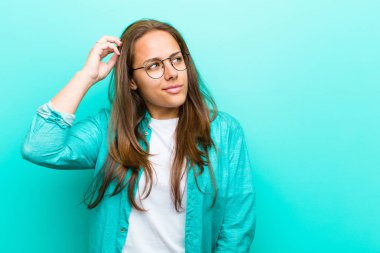 Genç kadın şaşkın ve kafası karışmış hissediyor, kafasını kaşıyor ve mavi arka plana bakıyor.