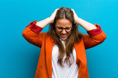 Genç bir kadın kendini stresli ve hayal kırıklığına uğramış hissediyor, el ele tutuşuyor, yorgun, mutsuz ve mavi arka planda migreni var.