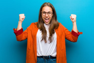 Genç kadın mutlu, pozitif ve başarılı hissediyor, zaferi kutluyor, başarıları kutluyor veya mavi arka plana karşı iyi şanslar diliyor.