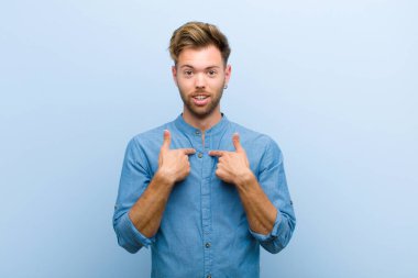 Genç işadamı mutlu, şaşkın ve gururlu hissediyor, mavi arka plana karşı heyecanlı ve şaşkın bir bakışla kendini işaret ediyor.