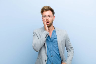 Genç işadamı şok olmuş ve şaşkın hissediyor. Mavi arka planda ağzı tamamen açık bir şekilde yüz yüze inanmamakta zorlanıyor.