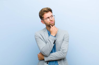 Genç işadamı düşünüyor, kuşkulu ve şaşkın hissediyor, farklı seçenekleri var, mavi arka plana karşı hangi kararı vermemiz gerektiğini merak ediyor.