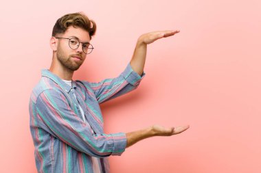 Genç bir adam iki eli yanda olan bir nesneyi tutuyor. Fotokopi alanı, pembe arka plana karşı bir nesneyi gösteriyor, sunuyor ya da reklamını yapıyor.