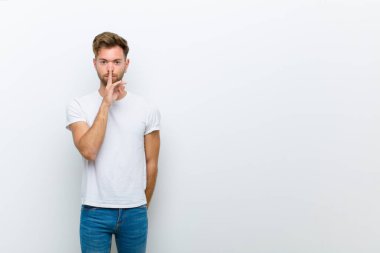 Genç adam ciddi görünüyor ve parmaklarını dudaklarına bastırarak sessizlik ya da sessizlik talep ediyor, beyaz arka plana karşı bir sır saklıyor.