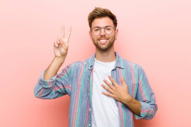 Genç adam mutlu, kendinden emin ve güvenilir görünüyor. Gülümsüyor ve zafer işareti gösteriyor. Pembe arka plana karşı olumlu bir tutum sergiliyor.