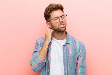 Genç adam kendini stresli, hayal kırıklığına uğramış ve yorgun hissediyor. Pembe arka planda endişeli ve sorunlu bir bakışla boynunu okşuyor.