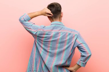 Genç adam bilgisiz ve kafası karışık, bir çözüm düşünüyor, eli kalçasında, diğeri kafasında, pembe arka planda.