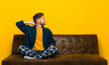 Genç adam pijama giyiyor, düşünüyor ya da şüphe ediyor, kafasını kaşıyor, kafası karışık ve kafası karışık hissediyor, geriye ya da arkaya bakıyor. Kanepede oturuyor.