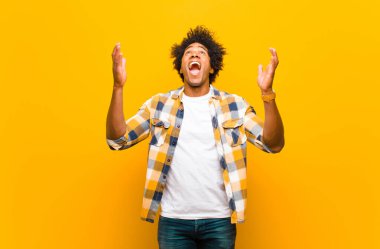 Genç siyah adam mutlu, şaşkın, şanslı ve şaşırmış hissediyor, zaferi kutluyor, iki eli de havada turuncu duvara karşı.