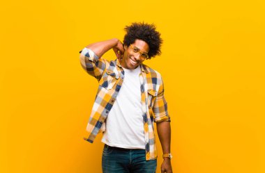 Genç siyah adam neşeli ve kendinden emin bir şekilde gülüyor. Turuncu duvarın karşısında mutlu, samimi bir gülümsemeyle.