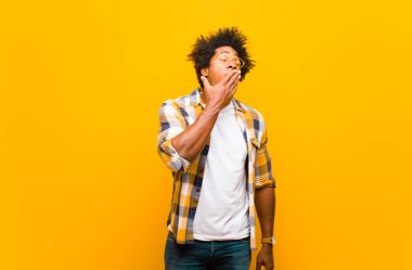 Genç siyah adam sabahın erken saatlerinde tembelce esniyor, uykulu görünüyor, yorgun ve turuncu duvara karşı sıkılmış.