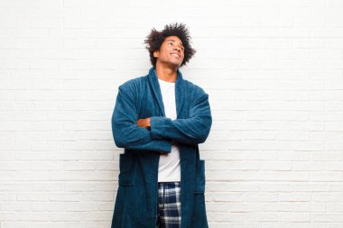 Genç siyah adam pijama giyiyor, mutlu, gururlu ve umutlu, merak ediyor ya da düşünüyor, tuğladan duvara yaslanmış kollarıyla fotokopi çekmeye bakıyor.