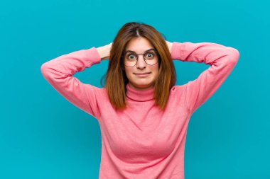 Genç, güzel bir kadın kendini stresli, endişeli, endişeli ya da korkmuş hissediyor, elleri başının üstünde, mavi arka planda hata yaparken panikliyor.