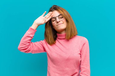 Genç ve güzel bir kadın stresli görünüyor, yorgun ve hayal kırıklığına uğramış, alnından ter damlıyor, mavi arka planda umutsuz ve bitkin hissediyor.