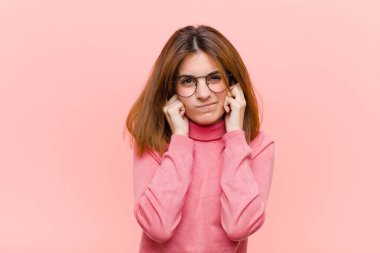 Genç, güzel bir kadın kızgın, stresli ve sinirli görünüyor, her iki kulağı da sağır edici bir ses, ses veya yüksek sesle kapatıyor pembe arka plana karşı müzik