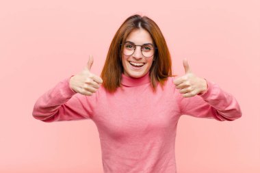 Genç ve güzel bir kadın gülümsüyor. Mutlu, pozitif, kendinden emin ve başarılı görünüyor. İki başparmağı da pembe arka planda.