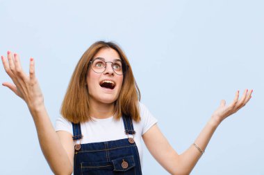 Genç, güzel bir kadın kendini mutlu, şaşkın, şanslı ve şaşırmış hissediyor. Zaferi kutlarken iki elini de havaya kaldırıp mavi arka plana yaslanıyor.