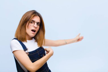 Genç, güzel bir kadın şaşkın ve şaşırmış hissediyor. Mavi arka planda şaşkınlık dolu, açık ağızlı bir bakışla fotokopi alanını işaret ediyor.