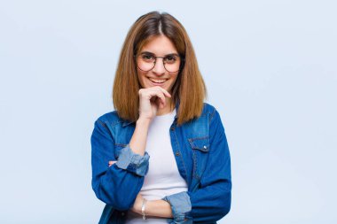 Genç ve güzel bir kadın mutlu görünüyor ve gülümsüyor, elleri çenesinde, merak ediyor ya da soru soruyor, mavi arka planla seçenekleri karşılaştırıyor.