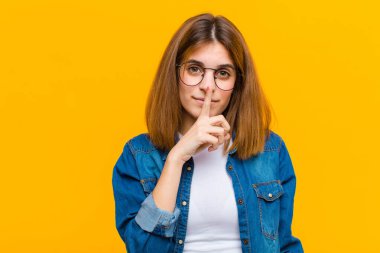 Genç ve güzel bir kadın ciddi görünüyor ve parmaklarını dudaklarına bastırıp sessizlik ya da sessizlik talep ediyor sarı arka plana karşı bir sır saklıyor.