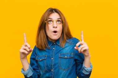 Genç ve güzel bir kadın korkuyla ve ağzı açık bir şekilde sarı arka planda şaşkın ve şaşkın bir bakışla yukarıya bakıyor.